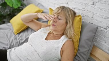 Stresli, sarışın, orta yaşlı bir kadın yatakta yatıyor. Endişeli bir şekilde, yatak odasının hücre hapsinde duygusal bir mücadeleyle karşı karşıya..