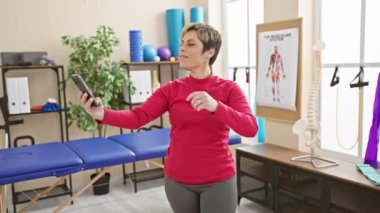 Klinikte gülümseyen bir kadın rehabilitasyon terapi odasında uzanırken akıllı telefonuyla selfie çekiyor..