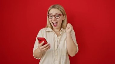 Heyecanlı genç sarışın kadın akıllı telefon karşısında büyük zaferi kutluyor. Sevincini kollarını kaldırıp kırmızı arka planda tek başına duruyor..