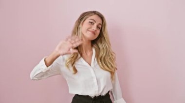 Tişörtlü neşeli sarışın kadın pembe izole edilmiş arka planda duruyor, kendine güvenerek el sallıyor, neşeli bir sırıtışla selam veriyor, olumlu duygular besliyor, gülümsüyor.