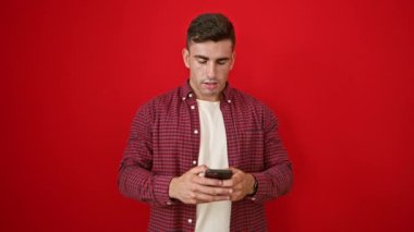 Gülümseyen, İspanyol asıllı genç adam mutluluk saçar, telefonu tutar ve kullanır. İzole edilmiş kırmızı duvardan mesaj atan, dijital sohbetin tadını çıkaran neşeli adamın portresi..