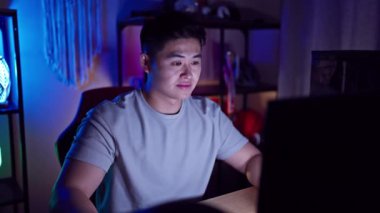 Neşeli genç Çinli erkek flamalı, kendine güvenen dişlek gülümsemeli, geceleri oyun odasında otururken mutlu bir şekilde video oyunu oynayan.