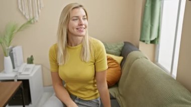 Neşeli genç sarışın kadın evde dostça bir merhaba sallıyor, sıradan bir tişörtle bir kulağından diğerine sırıtıyor, neşe ve güven saçıyor..