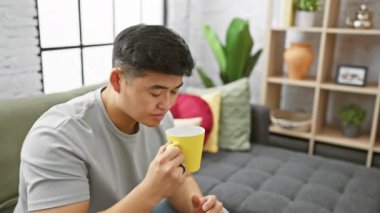Gündelik tişörtlü genç bir Asyalı modern bir apartman dairesinde, seçici dekorlu sarı bardaktan yudumluyor..