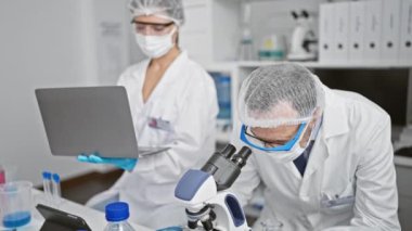 Laboratuvarda, 19 araştırmanın ortasında mikroskoplarıyla yaklaşan ve tıbbi maske takan iki bilimadamı birlikte.