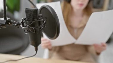 Beyaz kadın radyo stüdyosunda mikrofon ve pop filtresiyle senaryo okuyor, profesyonel bir kayıt seansını canlandırıyor..