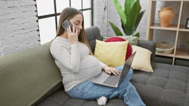 Modern bir oturma odasında dizüstü bilgisayar ve telefon kullanan İspanyol hamile kadın, evde annelik ve teknoloji kullanımı sergiliyor..