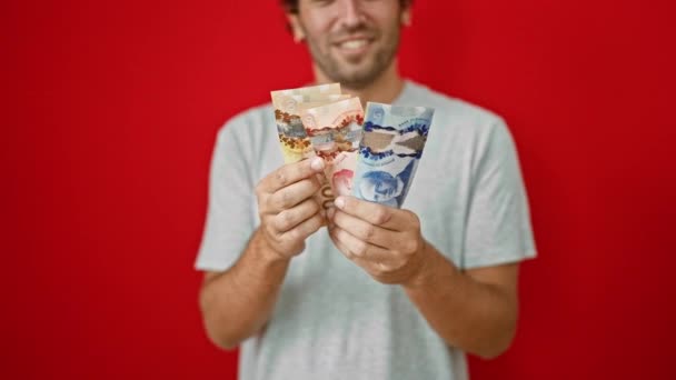 笑顔の若い男は 活気に満ちた赤い背景に孤立したカナダドル紙幣の山を喜んで数えています — ストック動画
