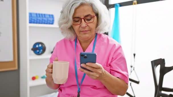 Felnőtt Rózsaszín Köpenyben Kávét Kortyolgat Ellenőrzi Telefonját Egy Klinika Pihenőjében — Stock videók