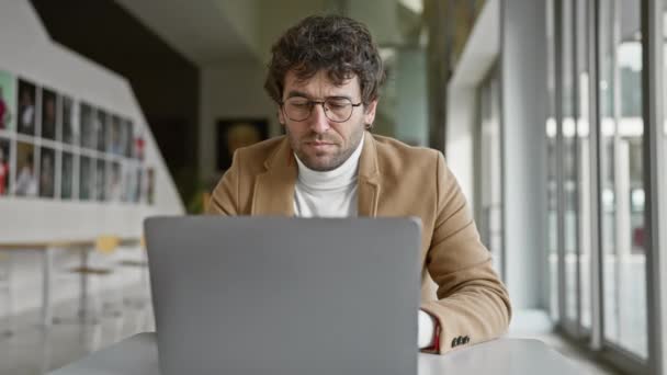 メガネとひげのあるヒスパニックな男性が 現代的なオフィス環境でラップトップに取り組んでいます — ストック動画