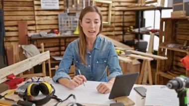 Beyaz kadın bir marangozhanede aletler, tablet ve güvenlik malzemeleriyle tasarımlar çiziyor..