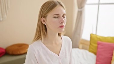 Beyaz kadın, rahat bir yatak odasında, renkli yastıklar ve doğal ışıkla huzur içinde meditasyon yapıyor..