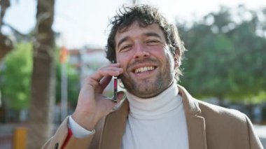 Gülümseyen, sakallı ve ceketli genç bir İspanyol güneşli bir günde telefonda konuşuyor..