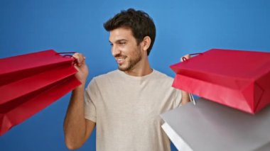 Sakallı yakışıklı genç adam mavi arka planda elinde renkli alışveriş torbalarıyla gülümsüyor.