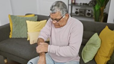 Gözlüklü, pembe kazaklı, gri bir kanepede oturan, koluna bakan son sınıf öğrencisi..