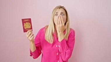 Şok olmuş genç sarışın kadın elleriyle ağzını kapatmaktan korkuyor, Kuzey İrlanda pasaportu ortaya çıktı! Şaşırmış ifade yüzünü soyutlanmış pembe arka planda dolduruyor.