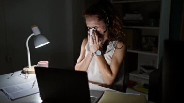 Geceleri dağınık ofis masasına hapşıran bir kadın. Masa lambası ve laptop ekranıyla aydınlatılıyor..