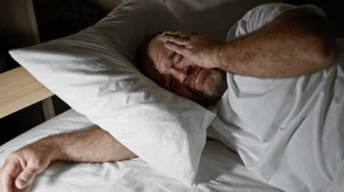 Orta yaşlı, sakallı, yorgun ve stresli beyaz bir adam loş ışıklı bir yatak odasında yatağa uzanmış, hayatın bir portresini gözler önüne seriyor.