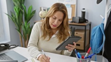 Ofiste çalışan odaklanmış Latin bir kadın elinde tablet tutarken notlar yazıyor..