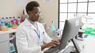 Laboratuvar önlüklü, modern bir laboratuvar ortamında bilgisayarla çalışan odaklanmış Afrikalı bir adam..