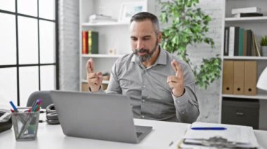 Ofiste sakallı, gri saçlı, neşeli, olgun bir adam. Modern, bitkilerle dolu bir işyerindeki dizüstü bilgisayarın içeriğine duygusal tepki veriyor..