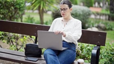 Genç, güzel, İspanyol bir kadın dizüstü bilgisayar kullanıyor, bankta oturuyor ve parkta işini bitiriyor.