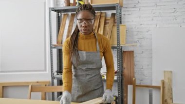 Afrikalı Amerikalı bir kadın, güvenlik gözlüğü ve gri önlük takarak, iyi donanımlı bir marangozluk atölyesinde kereste inceliyor..