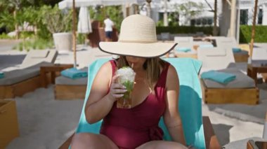 Gülümseyen bir kadın, geniş kenarlı bir şapkayla gölgelenmiş lüks bir sahil havuzunda mojito içmekten zevk alır..