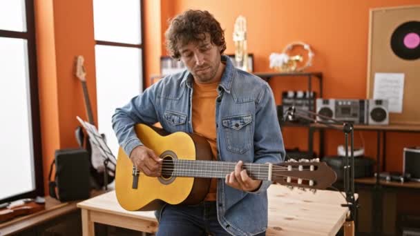 若いヒスパニックの男は 活気に満ちたオレンジ色の壁のある居心地の良い音楽スタジオでアコースティックギターを巧みに演奏します — ストック動画