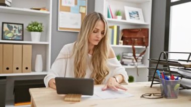 Odaklanmış sarışın bir kadın modern ofis ortamında arkaplanda raflar ve dekorlar olan belgeleri inceliyor..