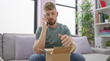Oturma odasında sakallı, düşünceli genç bir adam kucağında açık bir kutuyla telefonla konuşuyor..