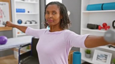 Orta yaşlı Afrikalı Amerikalı bir kadın rehabilitasyon kliniğinde dambıllarla fizik tedavi egzersizleri yapıyor.