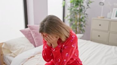 Üzüntülü bir kadın kalp desenli pijama giymiş, kapalı bir yatakta oturuyor, başını yatak odasında tutuyor..