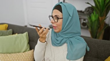 Başörtüsü takan olgun bir kadın rahat bir oturma odasında akıllı telefonuna sesli mesaj gönderiyor..