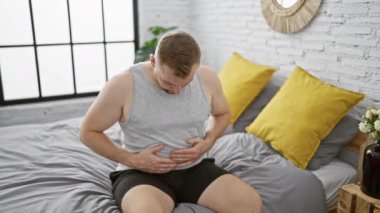 Kapalı bir yatakta oturan genç bir adam kederli bir ifadeyle karnına dokunuyor. Rahatsızlık ya da hastalığa işaret ediyor..