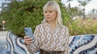Parkta otururken akıllı telefon kullanan, desenli kıyafetli, odaklanmış, sarışın bir kadın..