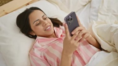 Yatak odasında akıllı telefon kullanırken gülümseyen İspanyol kadın evde rahatlık ve rahatlık tasvir ediyor.