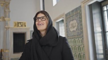 Gözlüklü ve eşarplı gülümseyen bir kadın süslü bir İstanbul sarayını ziyaret ediyor, turizm ve kültürü somutlaştırıyor..