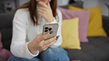 Beyaz kadın oturma odasında akıllı telefona göz atıyor, bağlantı ve modern yaşam tarzını resmediyor.