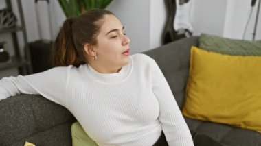 Genç bir İspanyol kadın, gri bir kanepenin üzerinde renkli yastıklarla rahatça uzanırken, elinde bir tüp ile evinde dinleniyor..