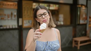 Neşeli İspanyol kadın Kyoto caddesinde nefis bir şekilde tatlı dondurmanın tadını çıkarıyor, güzel gülüşü bu Japon şehrinin geleneksel mimarisini deneyimlerken gözlüklerle çerçeveleniyor.