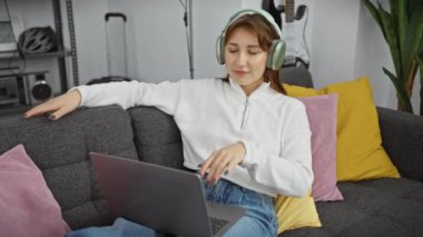 Oturma odasında dizüstü bilgisayar kullanırken kulaklıkla müzik dinleyen beyaz bir kadın..