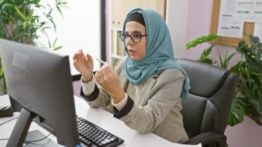 Gözlüklü olgun bir Müslüman kadın modern ofis ortamında bilgisayarının başında dikkatle çalışıyor..