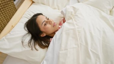 Genç bir İspanyol kadın düşünceli bir şekilde yatakta uzanıyor, rahat, kapalı bir yatak odası ortamında huzuru simgeliyor..