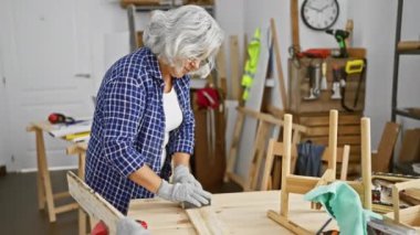 Eldiven giymiş olgun bir kadın marangozluk atölyesinde ahşap zımparalıyor..