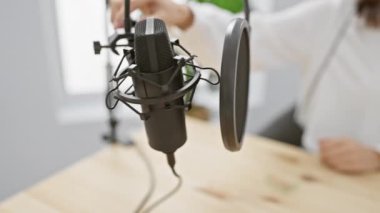 Beyaz bluzlu genç bir kadın, bitkilerle dolu parlak bir stüdyoda profesyonel mikrofon ve kulaklıkla ses kaydediyor.