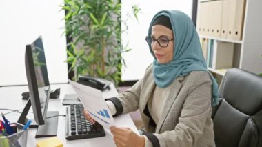Başörtülü profesyonel bir kadın ofis ortamında belgeleri analiz eder, verimliliği ve odaklanmayı betimler..