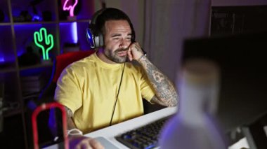 Sakallı İspanyol bir adam, neon ışıklı bir odada, kulaklık ve günlük kıyafetlerle oyun oynamaktan hoşlanır..