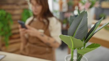 Gözlüklü ve dövmeli genç bir kadın canlı bir çiçekçide çalışırken akıllı telefon kullanıyor..