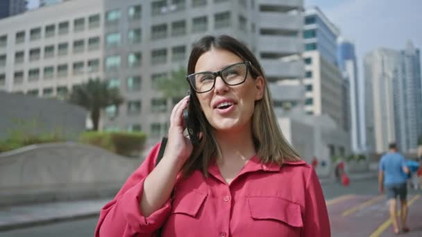 在现代摩天大楼的背景下 戴着眼镜在智能手机上说话的微笑的女人 — 图库视频影像